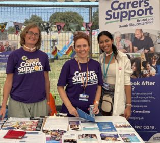Three Carers Support staff members stand behind an information table in the Community Area at Bristol Pride. From left to right are Lisa, Carmen and Ankita. The table is covered with leaflets, information packs and promotional materials about support