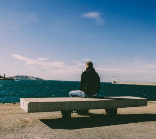 Person sitting alone on a concrete bench by a waterfront, looking out over a deep blue sea with distant hills and a lighthouse under a clear blue sky.