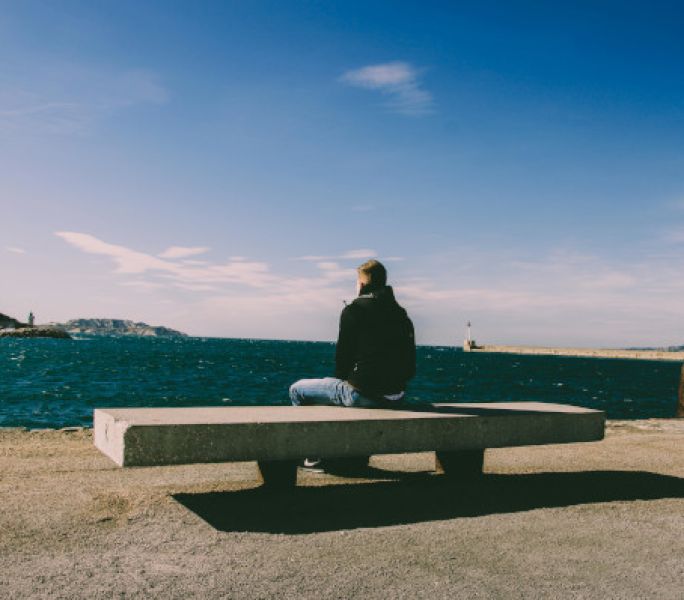 Person sitting alone on a concrete bench by a waterfront, looking out over a deep blue sea with distant hills and a lighthouse under a clear blue sky.