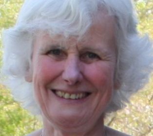 Head-and-shoulders portrait of Mary Whittington outdoors, with trees and greenery softly blurred in the background. Mary has short white hair and is facing the camera in natural daylight.