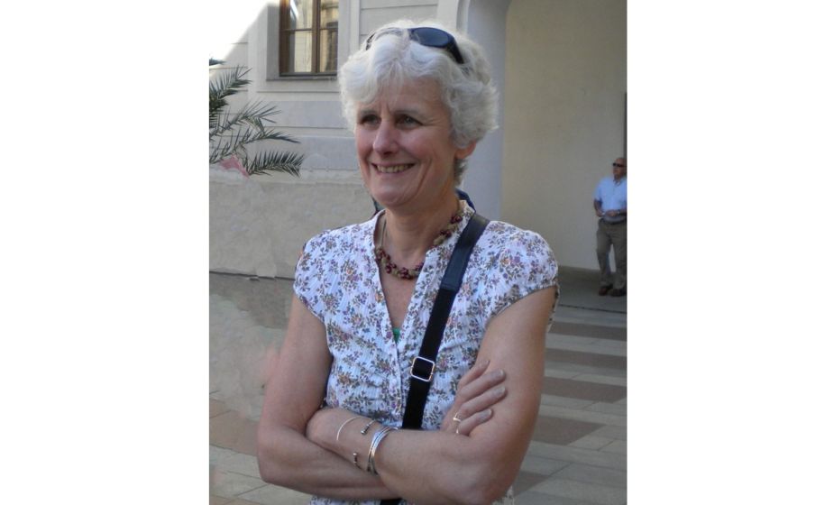 Copilot said: Alt text: Mary Whittington standing outdoors in a courtyard area, wearing a floral-patterned short-sleeved top and a necklace, with sunglasses resting on top of her head. Mary is standing with her arms folded, with a light-coloured buil