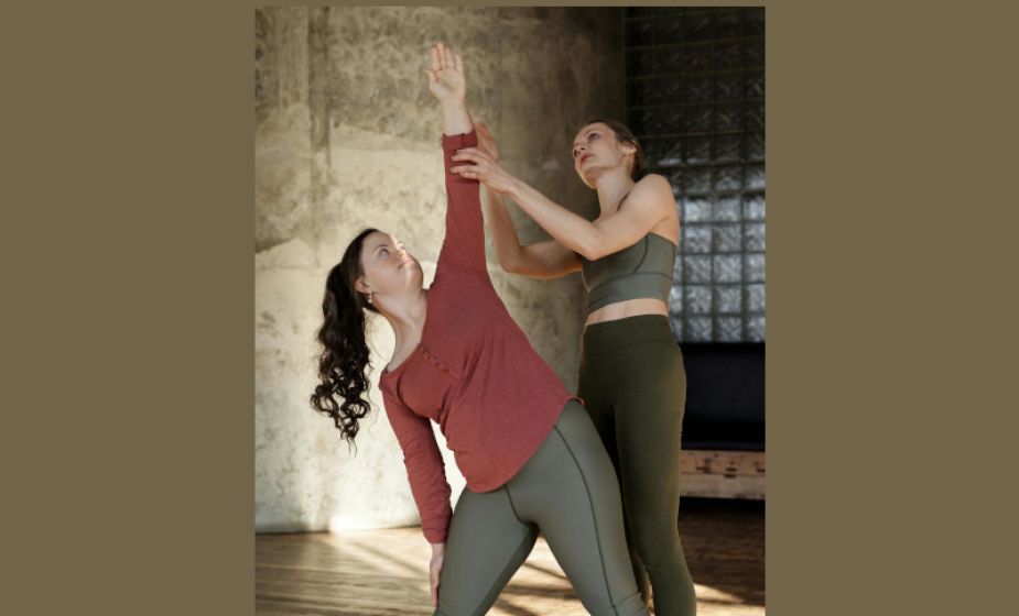 Two people taking part in a stretching or yoga session indoors, with one person supporting the other's posture during an exercise.