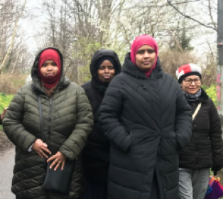 Five people wearing winter coats stand together on a tree-lined path in a park during a cold season.