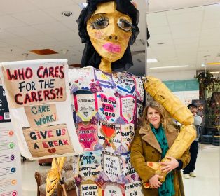 Large decorated puppet holding a sign that reads “Who Cares for the Carers?! Care Work Gives Us a Break”, standing beside a person in an indoor public space. The puppet is covered with colourful messages and illustrations about caring and support.