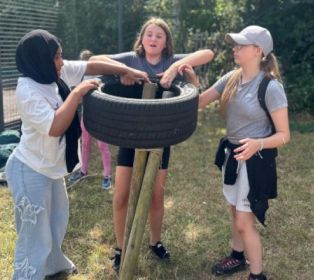 Three young carers work together on an outdoor team-building activity, lifting and positioning a large tyre around a set of wooden posts. The activity takes place on a grassy field, with other participants and trees visible in the background.