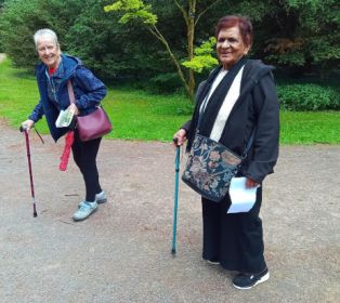 Two carers walk along a path at Westonbirt Arboretum during a Carers Support group visit. Both are using walking sticks and carrying event information as they walk through the arboretum's green woodland setting.