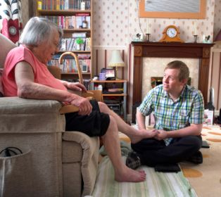 A carer sits on the floor of a living room, gently supporting the foot of an older person who is seated in an armchair. The room is homely, with bookshelves, a fireplace and patterned décor. The moment shows a quiet, caring interaction.