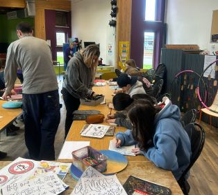 People taking part in a creative group activity in a community hall, seated around tables with colouring sheets, pencils and craft materials.