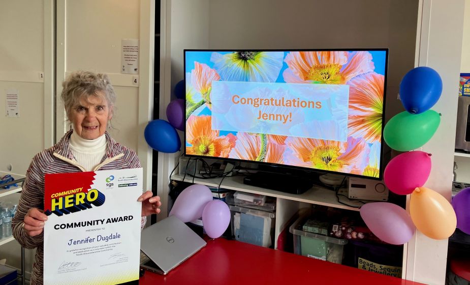Jenny Dugdale holding a Community Hero Community Award certificate in an indoor room decorated with colourful balloons. Behind the person, a television screen displays the message “Congratulations Jenny!” on a floral background. A red table, laptop a