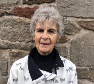 Jenny Dugdale standing in front of a stone wall, wearing a patterned light-coloured jacket over a dark high-neck top. The portrait-style photograph shows Jenny from the chest up against the textured stone background.