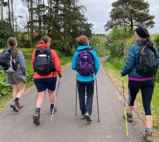 Four people walking along a country lane using Nordic walking poles. The group is viewed from behind, wearing outdoor clothing and backpacks. Trees, hedges and open countryside line the route under an overcast sky.
