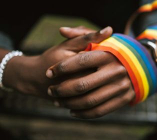 Close-up of two people holding hands, with one wrist wrapped in a rainbow Pride flag and rainbow-coloured wristbands visible. The image is softly focused in the background and symbolises support, connection and LGBTQIA+ commun
