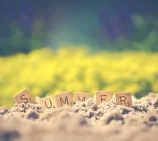 Wooden letter tiles spelling “SUMMER” partially buried in sand, with a blurred field of yellow flowers in the background and soft sunlight creating a warm, seasonal atmosphere.