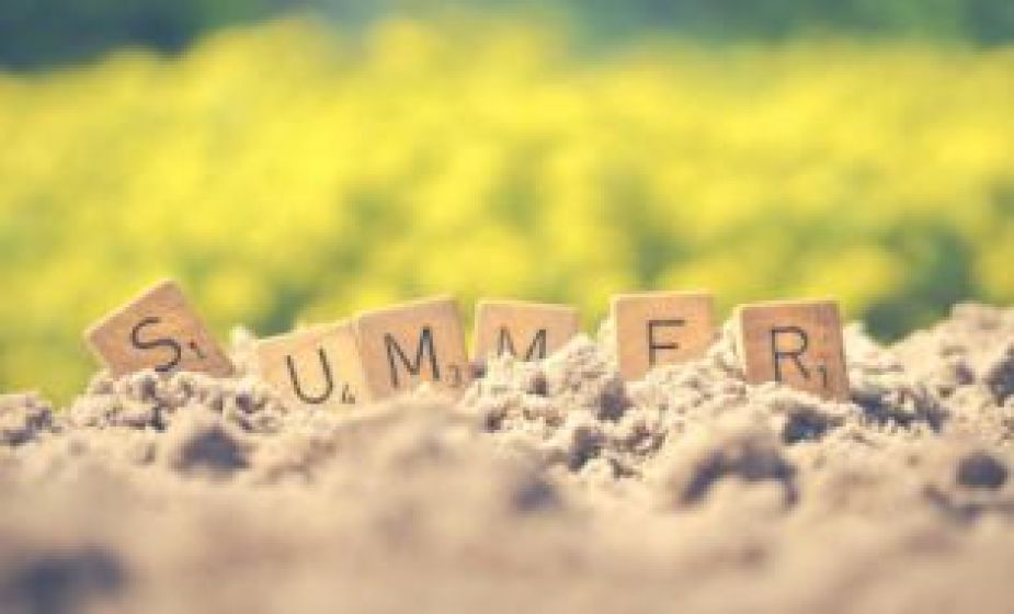 Wooden letter tiles spelling “SUMMER” partially buried in sand, with a blurred field of yellow flowers in the background and soft sunlight creating a warm, seasonal atmosphere.