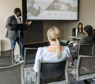 People seated in a meeting room watching a presentation, with a speaker standing at the front beside a projection screen.