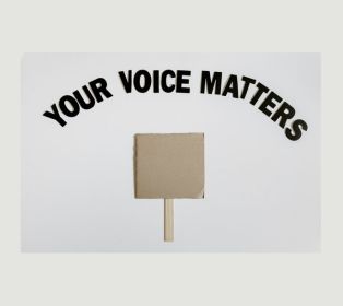 A blank cardboard placard on a wooden stick is positioned against a white background beneath the words “YOUR VOICE MATTERS” in large black letters. The simple graphic is designed to represent sharing opinions, feedback and having a say in decisions.