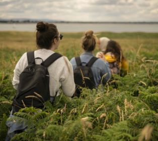 Three people sitting together in long grass and bracken near a coastal landscape. Two people are wearing backpacks, and one person is holding a small child. The group is viewed from behind, looking out across the open countrys