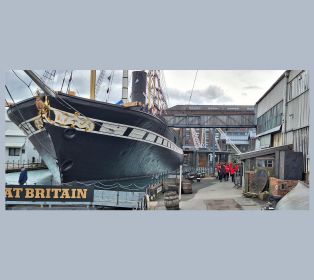 Black-hulled historic ship SS Great Britain docked in Bristol’s dry dock, with ornate gold detailing at the bow. A walkway runs alongside the ship past dockside buildings and barrels, where a group of visitors in red tops walk towards the ship.
