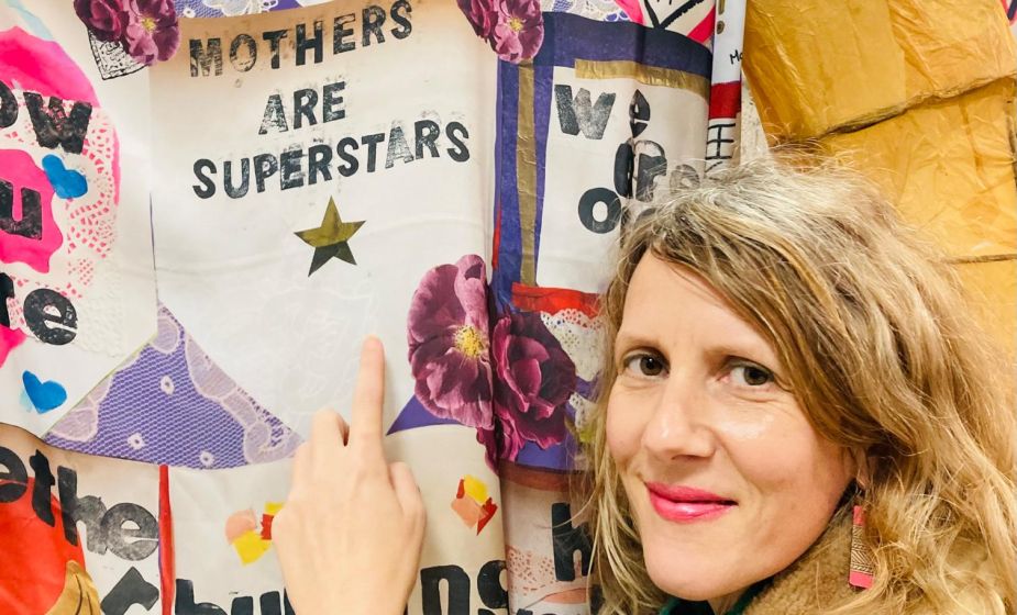 Dahlia standing beside a large textile artwork, pointing to a panel that reads “Mothers Are Superstars”, surrounded by decorative flowers, stars and other handmade messages.