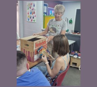 Volunteer leading a craft activity, holding an open cardboard box while participants sit around a table handling colourful materials in a community setting.