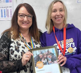 Two people standing indoors holding a framed certificate or award. One person is wearing a purple Young Carers Support hoodie and staff lanyard. The framed award includes a photograph of young carers and the words “Young Carers”. A whiteboard and edu