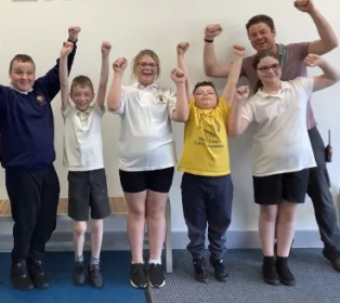 Five young people and one adult standing indoors with both arms raised above their heads. The group is arranged in a line against a plain wall, with most wearing school uniforms and one wearing a yellow T-shirt. The image captures a group activity or
