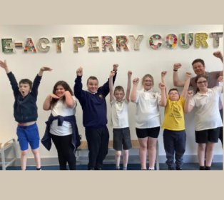 A group of school children and a teacher standing indoors with their arms raised in celebration, smiling beneath a wall sign that reads “E-ACT Perry Court.”