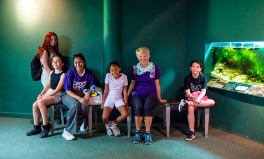 Five young carers and a Young Carers worker sit and stand on benches inside an indoor exhibit area at Slimbridge Wetland Centre. The group is gathered beside a large aquarium display filled with aquatic plants, with dark green walls forming the backg