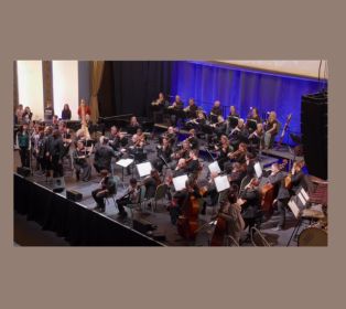 Bath Philharmonia Orchestra performing on stage during a concert. Musicians are seated in sections playing string and wind instruments, while a conductor stands at the front directing the performance. A choir is standing to one side of the stage, wit
