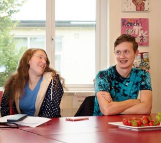 Two young carers seated at a red table in a bright room, with papers and a plate of strawberries in front of them.
